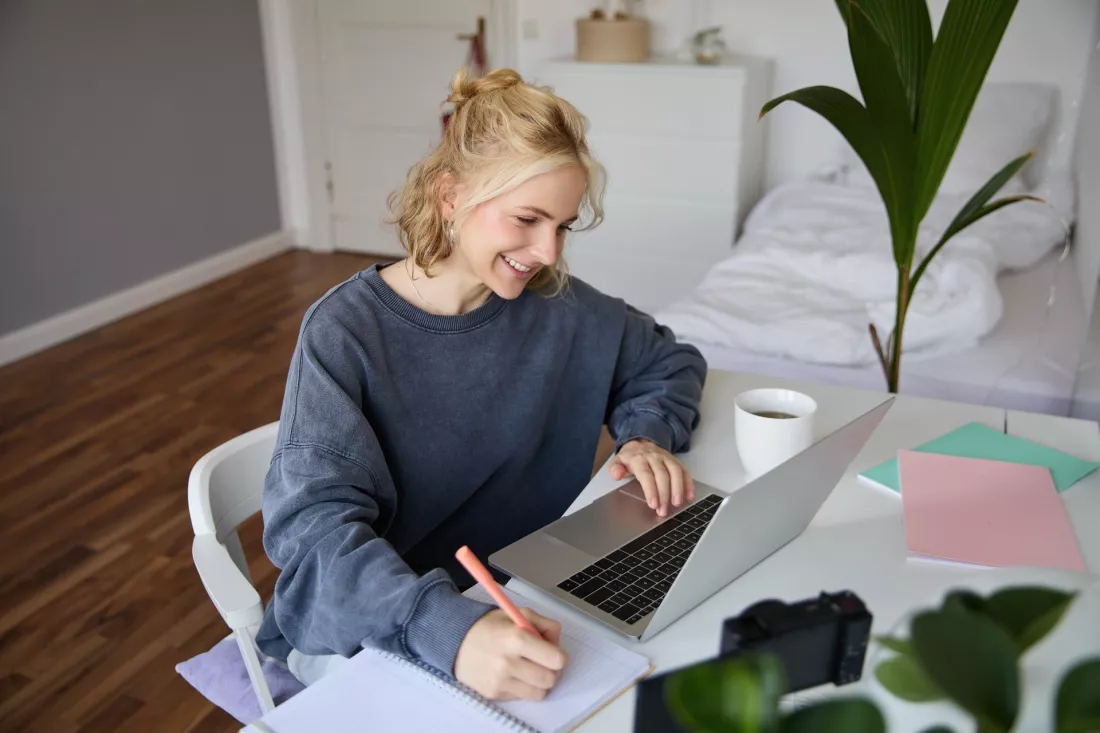 Uśmiechnięta studentka uczy się online przy laptopie i robi notatki