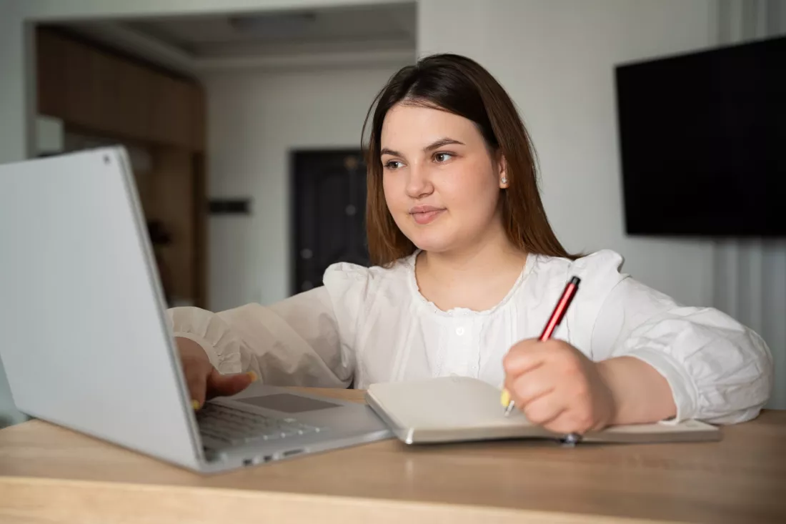 Mloda kobieta uczy sie online przy laptopie i robi notatki w domu