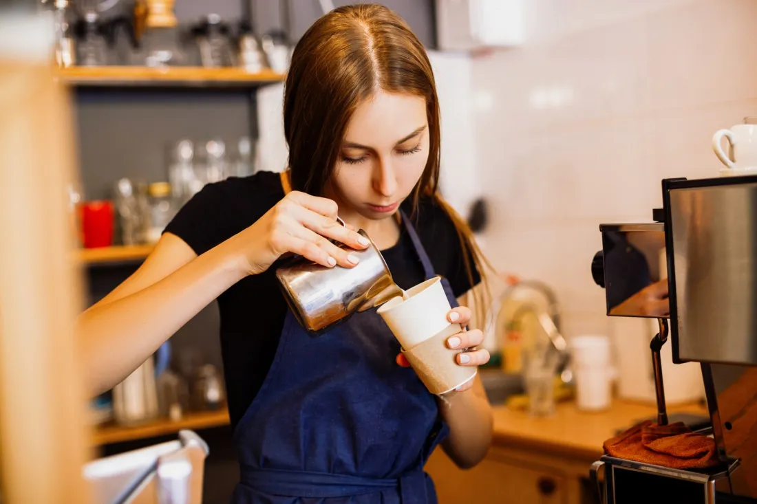 Młoda baristka nalewa mleko do kubka z kawą w kawiarni